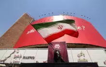 شخص يحمل العلم الإيراني أمام لوحة إعلانية ضخمة مناهضة للولايات المتحدة في ساحة "ولي عصر" بطهران في 19 يوليو/تموز
