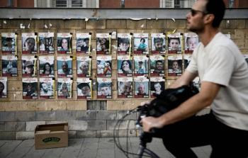 إسرائيلي يسير على دراجة هوائية أمام لافتة فيها صورا لأسرى محتجزين في غزة